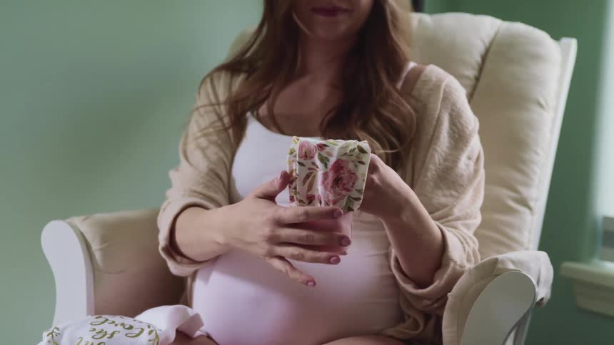 Pregnant female sits in rocking chair in a baby nursery and carefully folds a pair of pants for baby in half