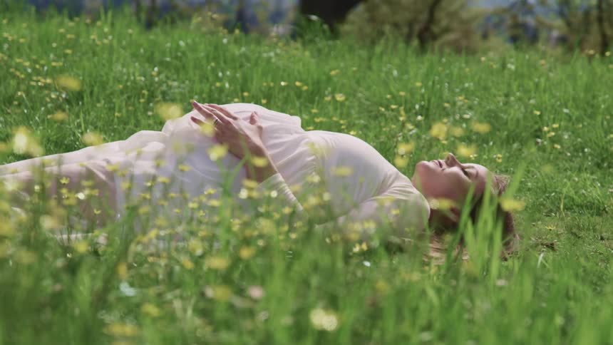 Pregnant woman with a white dress lies down on the green grass and holds her belly on a sunny day