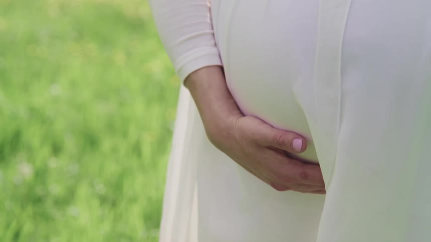 A pregnant woman with a white dress rubbing her stomach with grass in the background