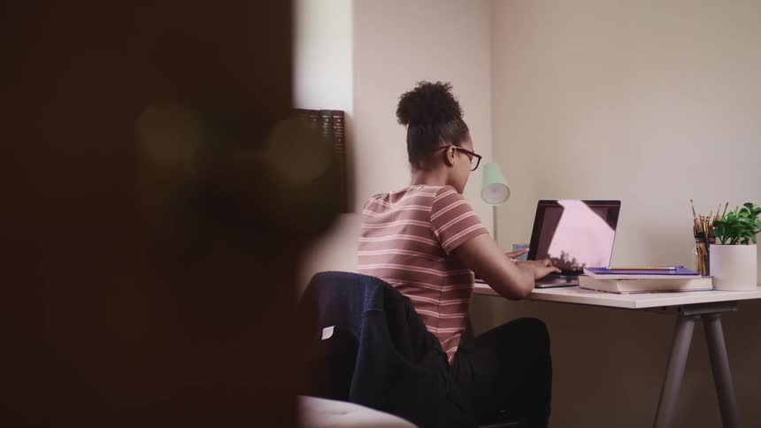Black woman with glasses and a striped t shirt sits at a desk and types on a laptop in a room