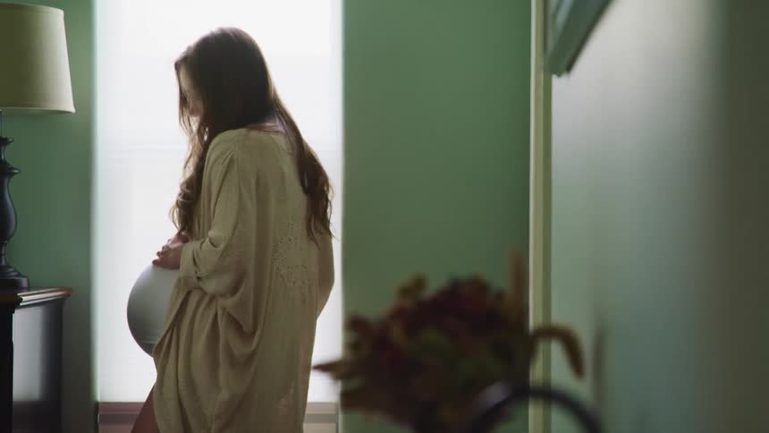 Pregnant woman with long brown curly hair touches her belly in a room at home