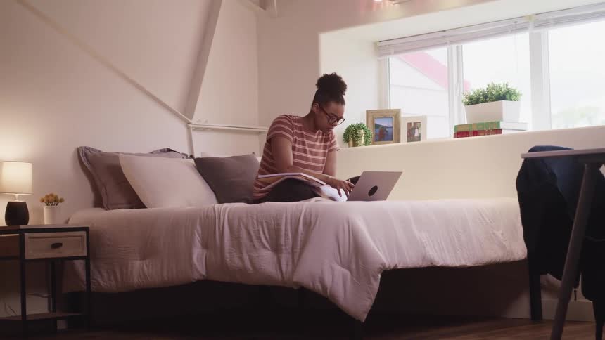 Black woman with glasses and a striped t shirt sits on a bed uses a laptop and writes in a notebook in the bedroom