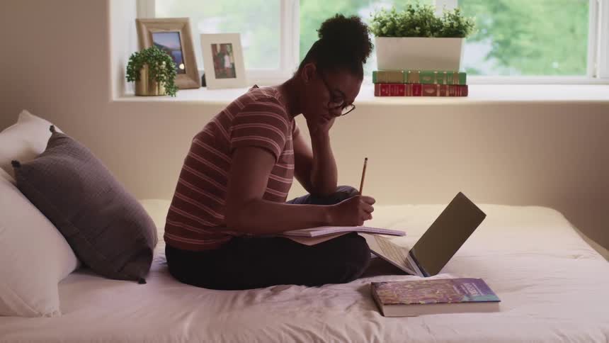 Black woman with glasses and striped shirt sits on a bed and takes notes while using a laptop in her room