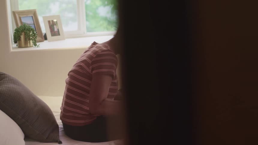Black woman with glasses and a striped t shirt sitting on a bed uses a laptop and writes in a notebook in her room