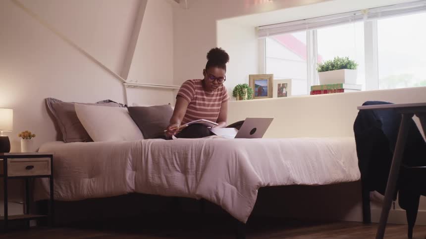 Black woman sits on a bed with a laptop as she looks through a book