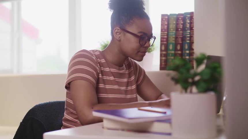 Black woman with glasses and striped t shirt sitting at a table uses a laptop in a room