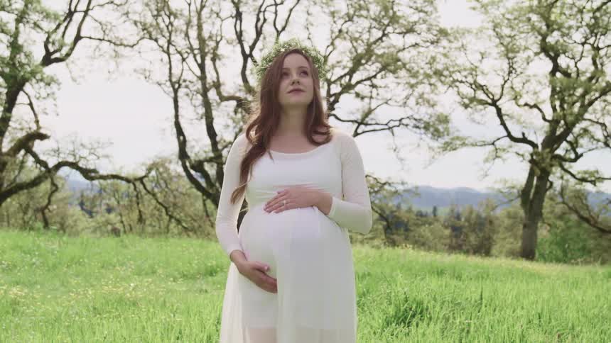 Pregnant woman with a white dress a flower crown and long brown curly hair stands on the grass and holds her belly