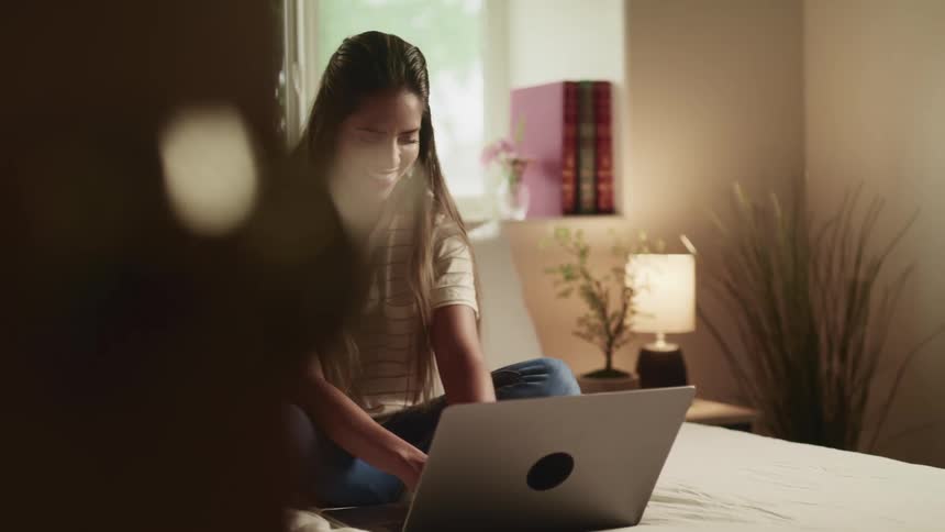 A woman sits cross legged on her bed as she smiles while using a laptop