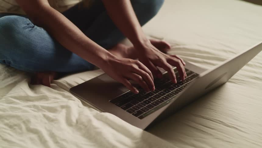 Females sitting on bed cross legged as she types on an open laptop in front of her