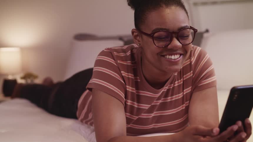 Black teenager lays on a bed and laughs as she waves to her cell phone