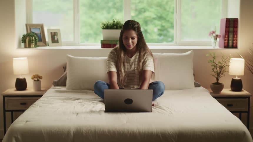 Brunette woman with brown hair a white t shirt and jeans sits on a bed and types on a laptop in the bedroom