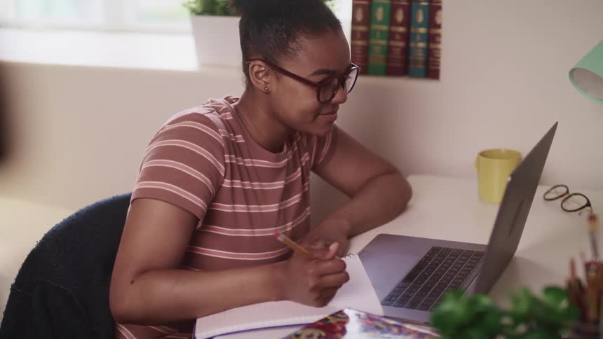 Black woman with glasses a striped shirt sits at a desk looks at a laptop and nods her head then takes a note in a room