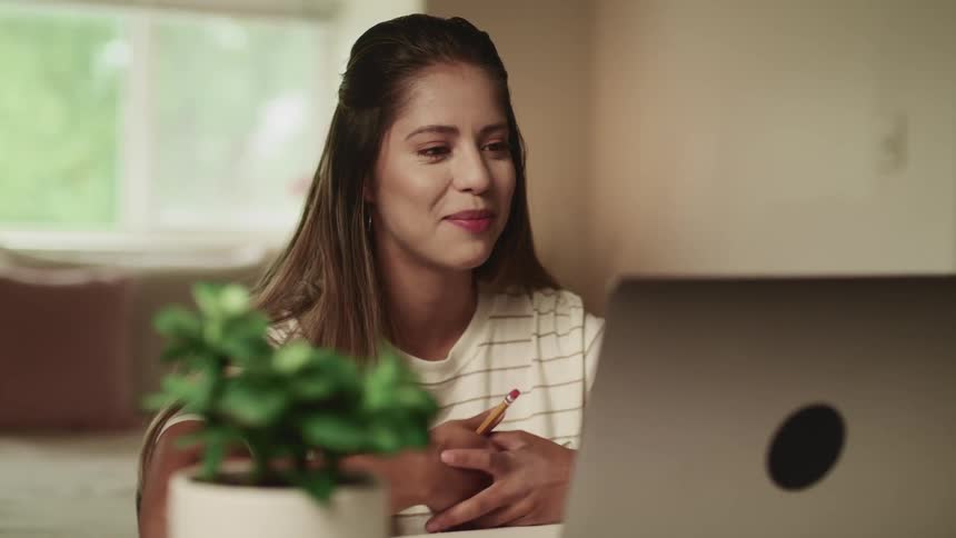 Female teenager waves and holds a pencil as she smiles and talks towards her laptop