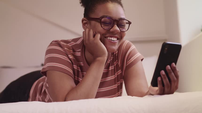 Young woman talks on her smartphone while laying on the bed in her bedroom at home