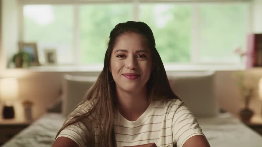 A woman sits at the end of her bed and smiles while nodding