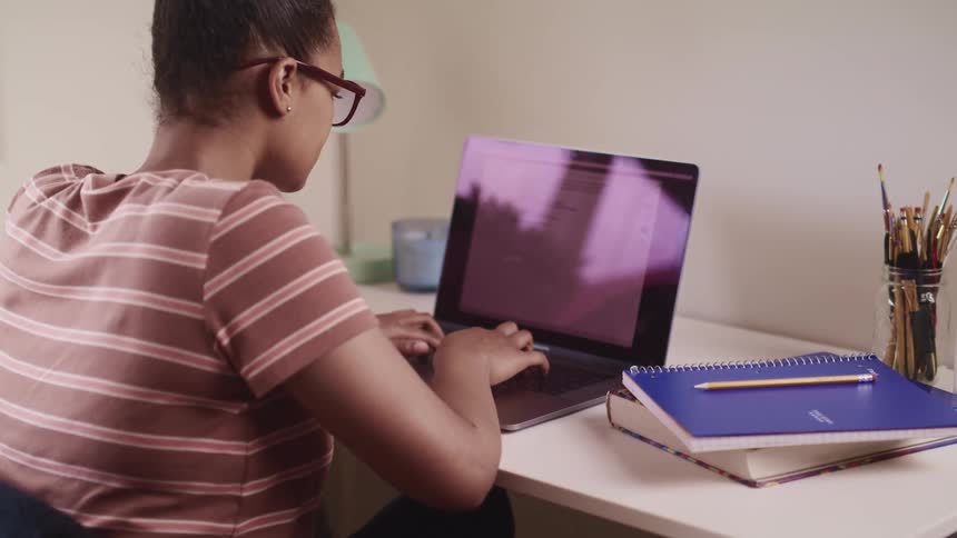 Black woman with glasses and striped t shirt sits at a desk and uses a laptop in a room
