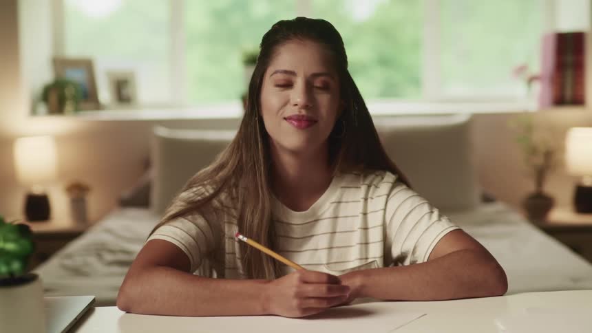 Brunette woman with a white t shirt sitting at a desk listens to the speaker and takes a note in an online lesson