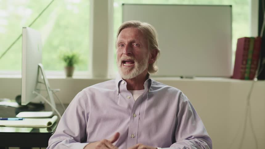 Middle aged man with a beard and a shirt sits in an armchair talks to the camera