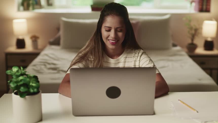 Female sitting at a desk in the bedroom smiles as she types and looks at the laptop screen