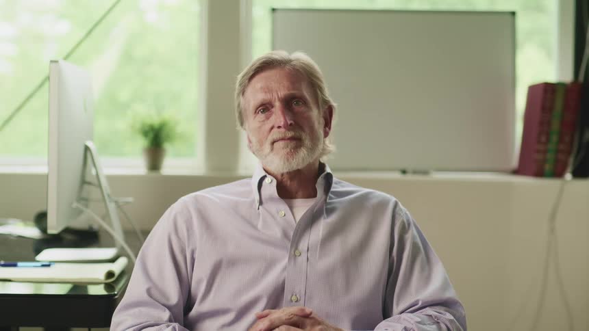A man laughs smiles and nods while looking into the camera from his seat in an office