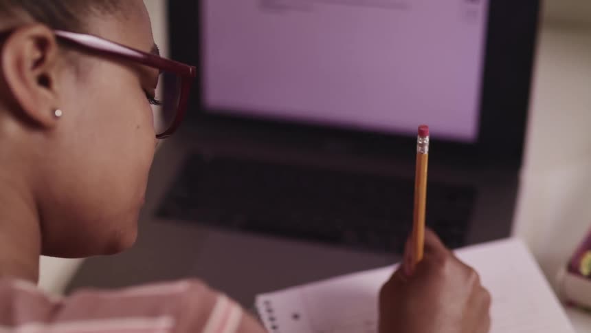 Female studies as she sits at her desk at home in front of a laptop and uses a pencil to write in a notebook