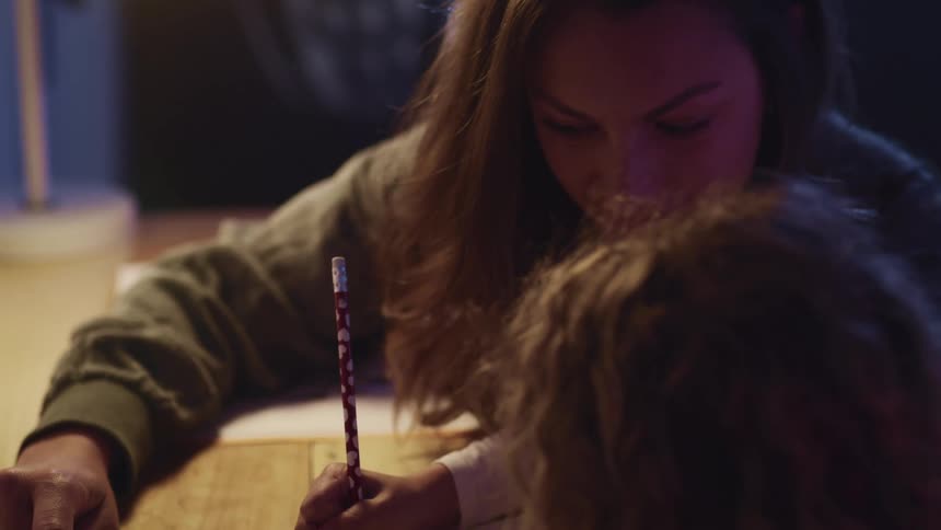 A woman watches as a child sitting next to her at a table uses a pencil to write on a piece of paper