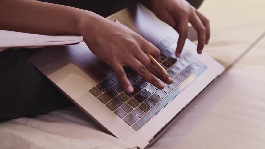 Black woman sitting on a bed holds a pencil and types on a laptop