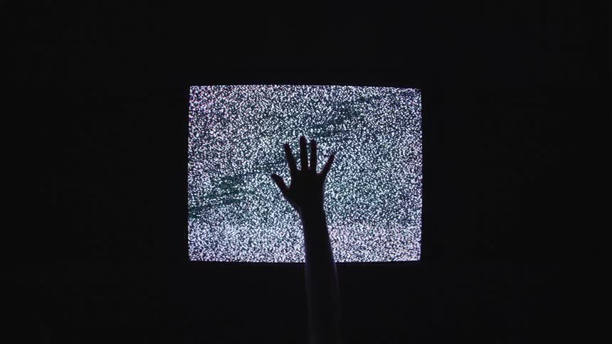 In a darkened room a small screen displays only static as someones hand reaches upwards in front of it