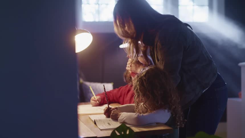 A woman gives a high five to one of a pair of little girls sitting at a desk writing with pencils