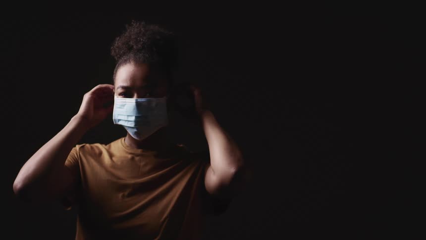 Black woman with a brown t shirt wears a light blue face mask on a dark background