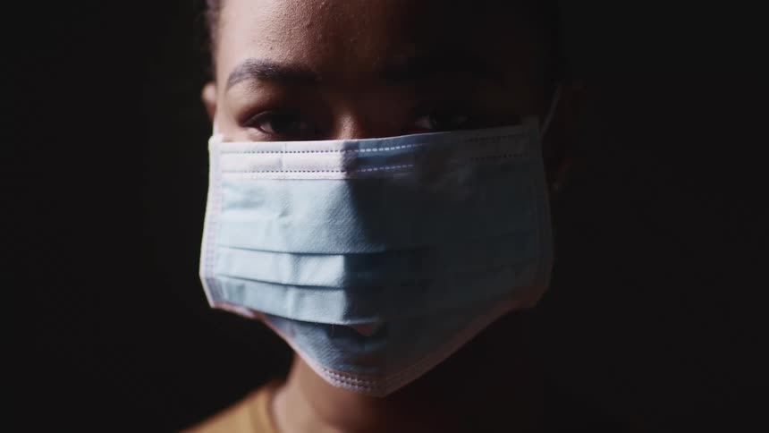 A young black woman in a medical mask looks into a camera while standing in a dark room