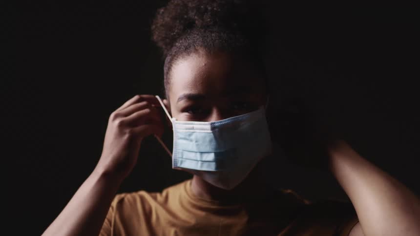 Black woman with a brown t shirt wears a light blue face mask on a dark background