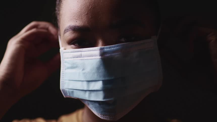 Black woman with a brown t shirt wears a light blue face mask on the dark background