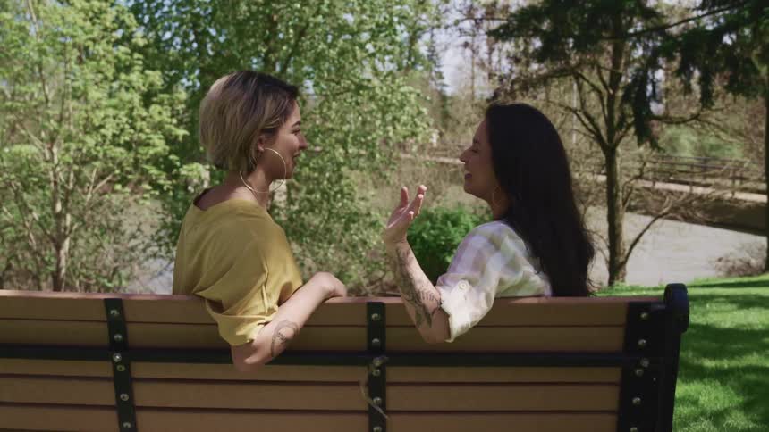 Woman with dirty blonde hair and hoop earrings and woman with dark hair and hoop earrings sits on a park bench in the public park