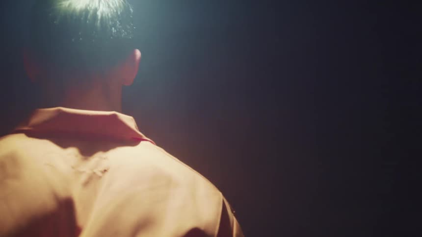 Man wearing orange shirt stands still under a swinging white light in a dark room