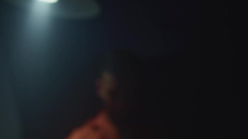 A young man without a shirt stands below a light that swings back and forth