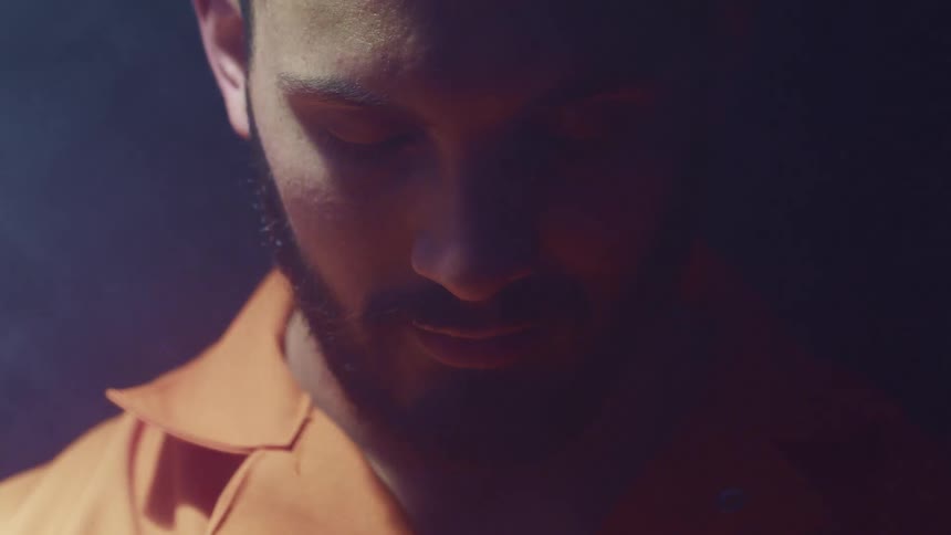 Bearded man wearing orange shirt in a dark room under swinging white light