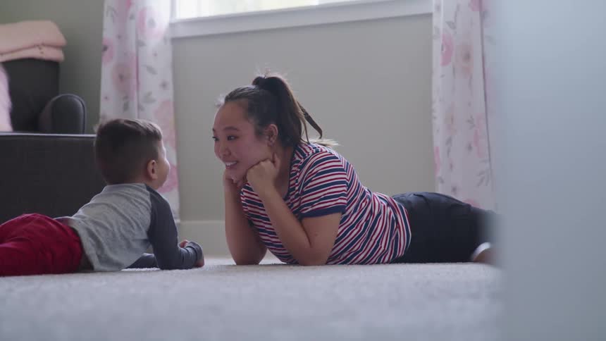 A woman playfully talks to child as the two lie facing each other on the carpeted floor of a living room