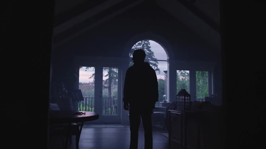 A man stands still while looking around a dim room illuminated onto by pale sunlight coming through the large windows
