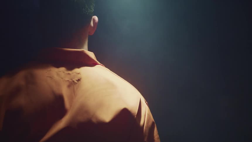 A man in an orange prison jumpsuit waits motionlessly under a swinging light in a dark room