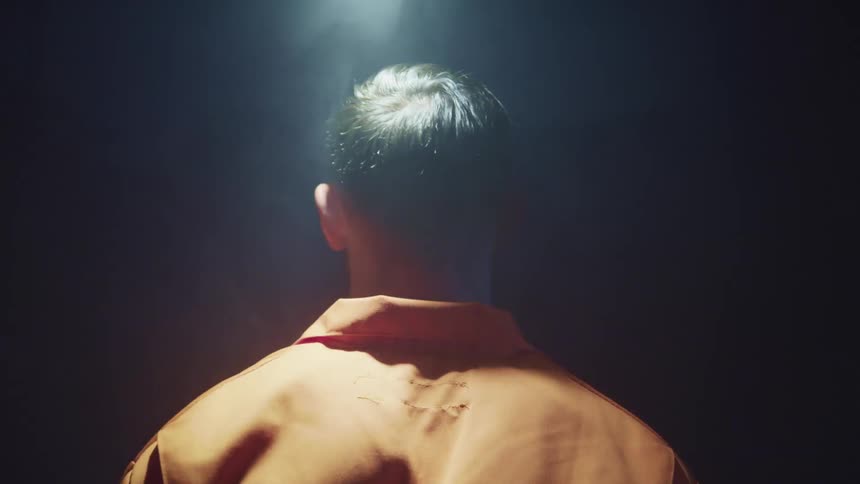 A man in an orange prison jumpsuit stands motionlessly under a swinging light in a dark room as a strobe light flashing on him from the side
