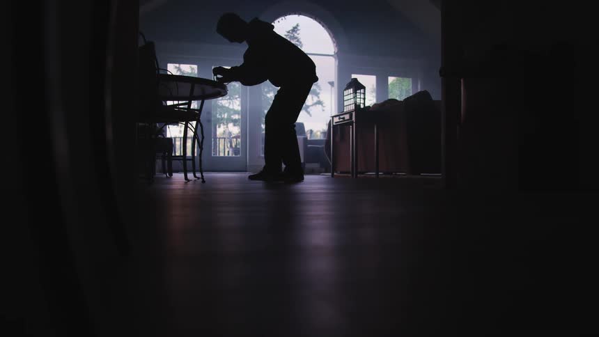 Person wearing hoodie quietly walks into dimly lit living room pocketing items on table before stealthily leaving