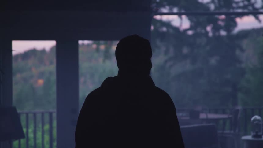 A man in a mask and dark clothing looks around a dimly lit living room