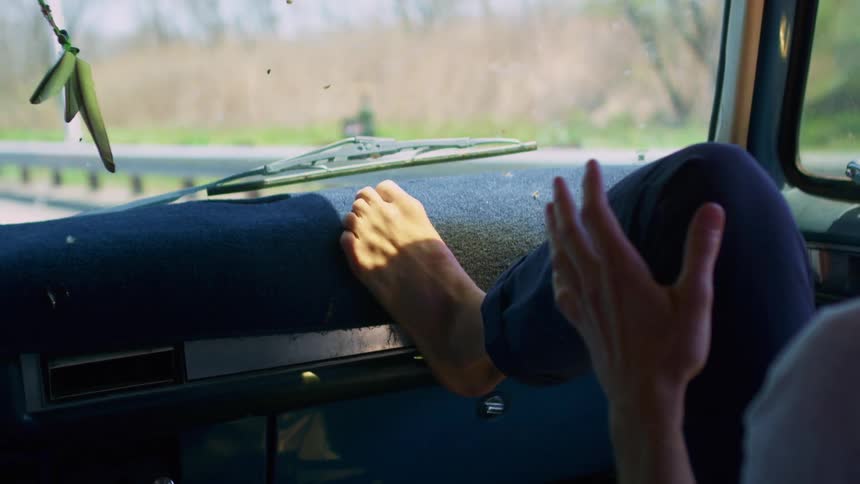 Male passenger leaning forward hits both hands on the dashboard then leans back in his seat while female driver seen in the rearview mirror smiles slightly and continues to drive along a road in the country