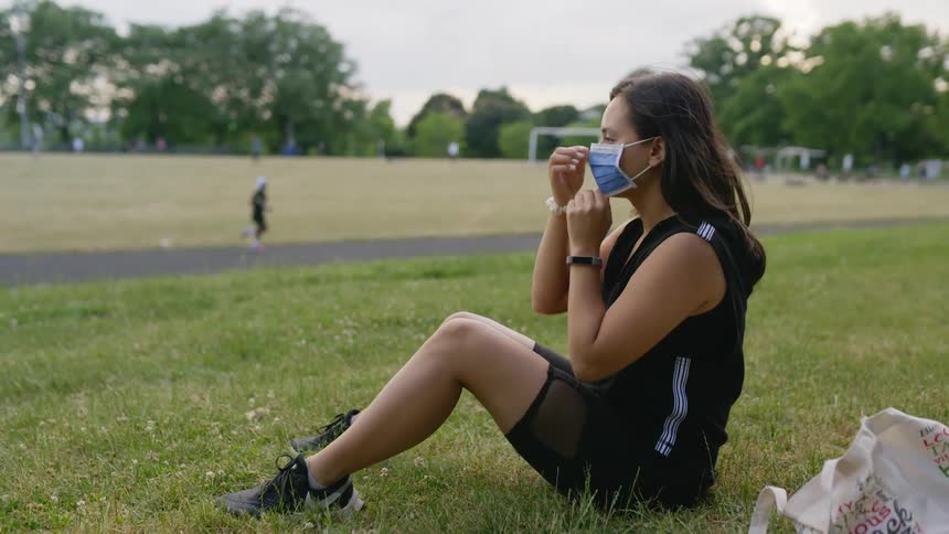 Woman sitting on grass dons face mask as jogger runsby on track