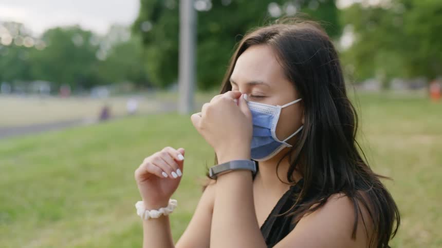 Brunette woman with dark hair sitting on grass fields wears a blue face mask in the public park