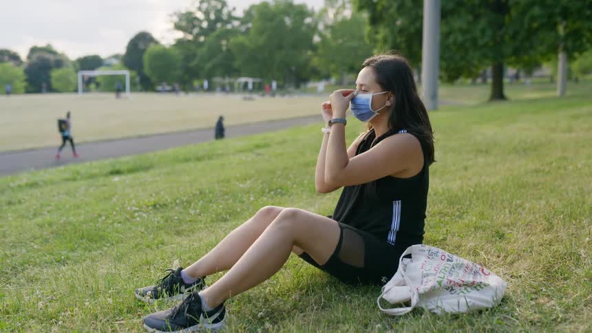 Woman sitting in the grass in a park takes a face mask from her purse and puts it on