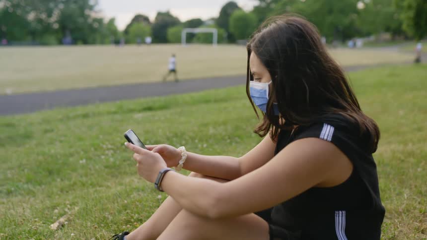 Woman wearing face mask sits on grass after texting