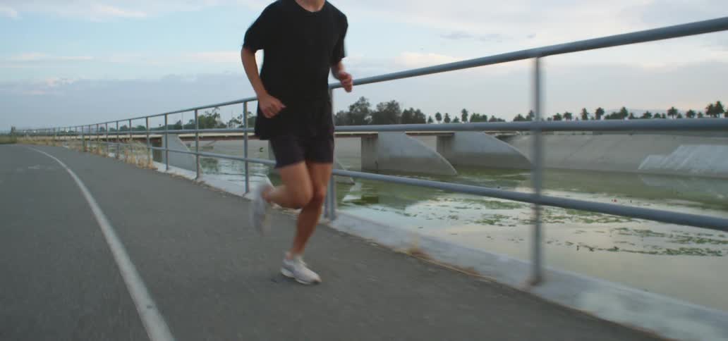 Man with a pair of white shoes black running shorts and a t shirt runs along an asphalt road on a cloudy day