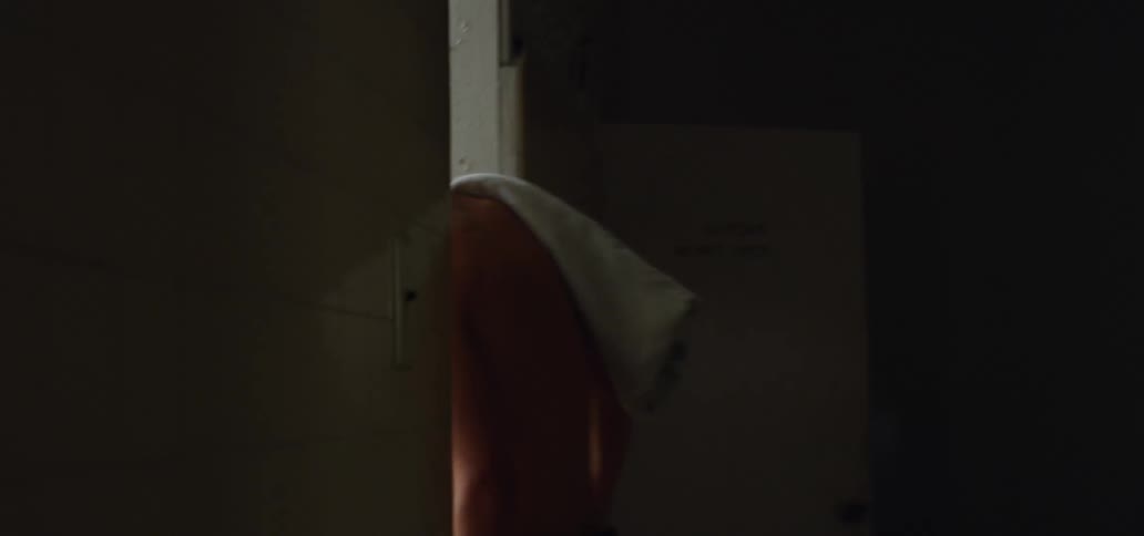 A shirtless young man with a towel on his shoulder wakes down a dark hallway before entering the bathroom